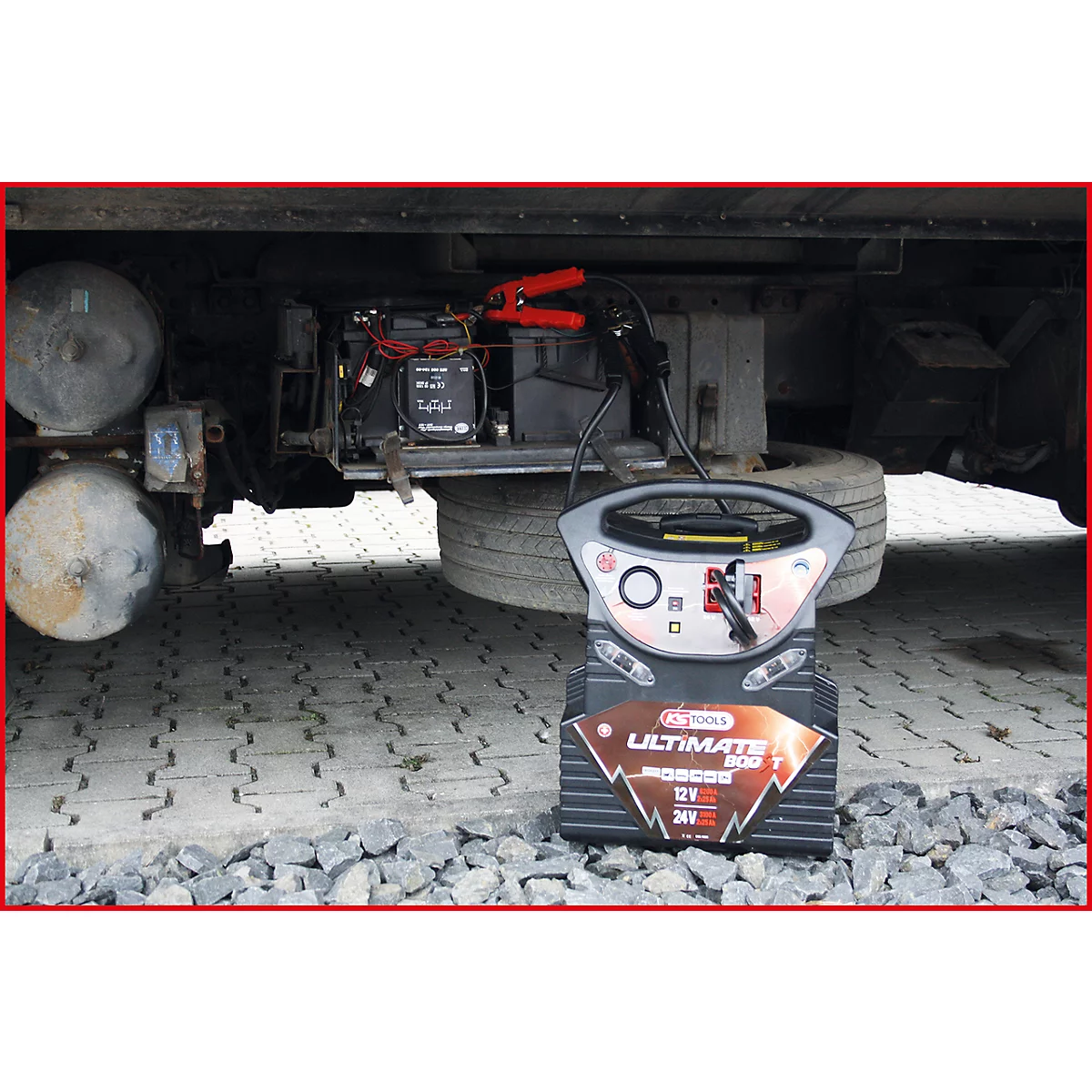 Ein Starthilfegerät steht vor einem LKW. Rote Kabelklemmen sind mit der Batterie verbunden.